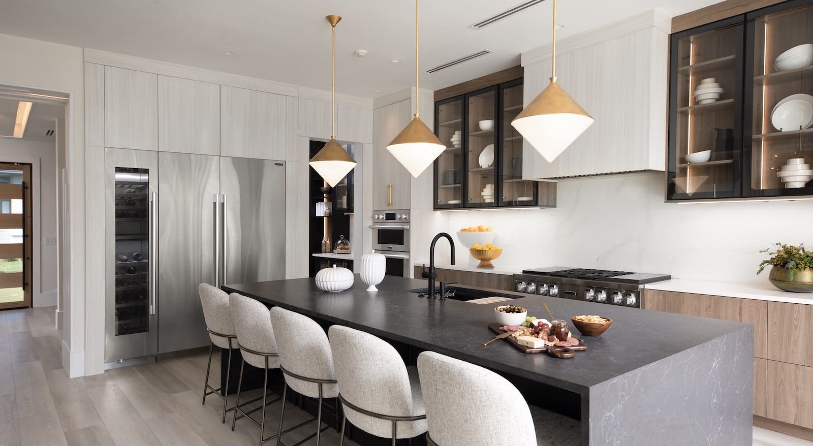A mix of textures, colors, and finishes in neutral yet warm tones creates a serene, inviting ambience in the home&rsquo;s main kitchen. The center island&rsquo;s dark-toned quartz surface contrasts with the light-toned flooring, while dramatic pendant lights provide a focal point for the space.
