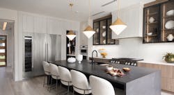 A mix of textures, colors, and finishes in neutral yet warm tones creates a serene, inviting ambience in the home’s main kitchen. The center island’s dark-toned quartz surface contrasts with the light-toned flooring, while dramatic pendant lights provide a focal point for the space. A mix of textures, colors, and finishes in neutral yet warm tones creates a serene, inviting ambience in the home’s main kitchen. The center island’s dark-toned quartz surface contrasts with the light-toned flooring, while dramatic pendant lights provide a focal point for the space.