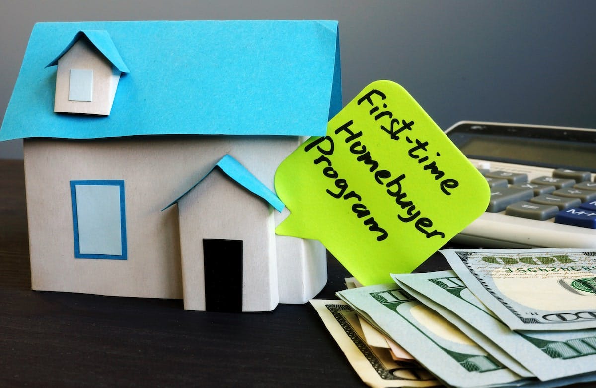 Pile of U.S. currency next to paper house with post-in note reading 'First-Time Homebuyer Program'
