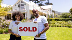 A couple stands in front of their newly purchased home, smiling and holding up a sign that reads 'Sold.'