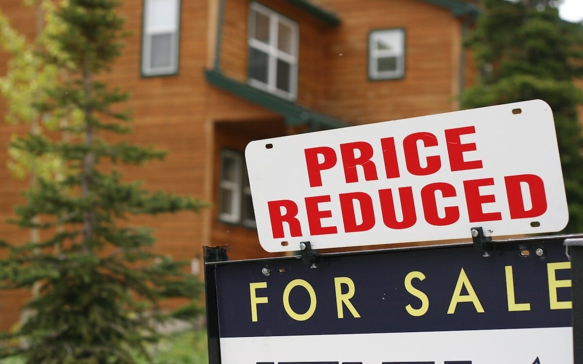 A 'For Sale' sign with a 'Price Reduced' sign added above it sit in front of an older home