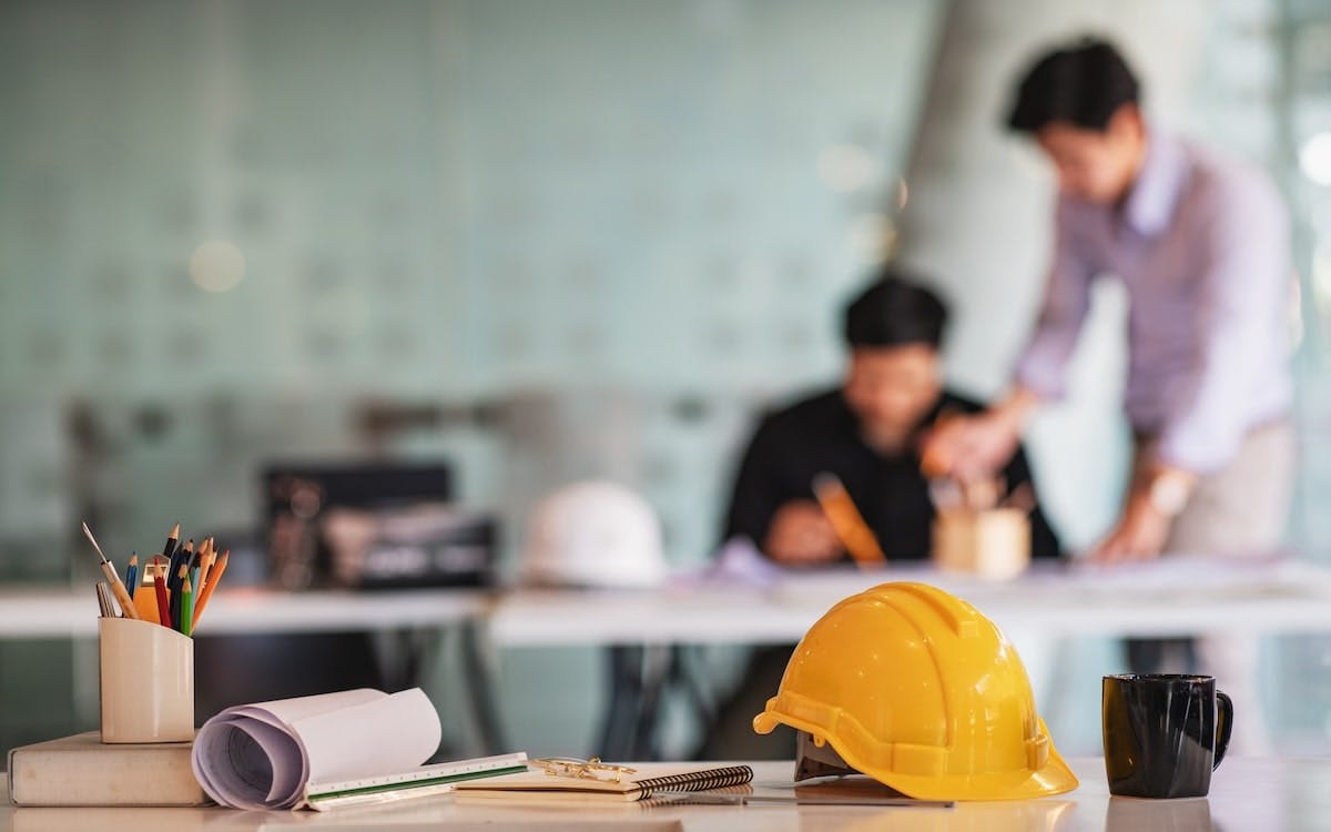 A student and teacher go over an assignment in the background. In the foreground, there is a table with some papers and a hard hat.