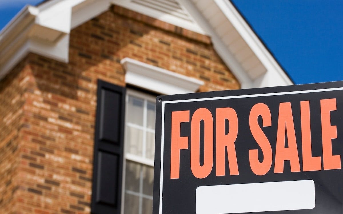 A 'For Sale' sign sits in the front yard of a brick single-family home