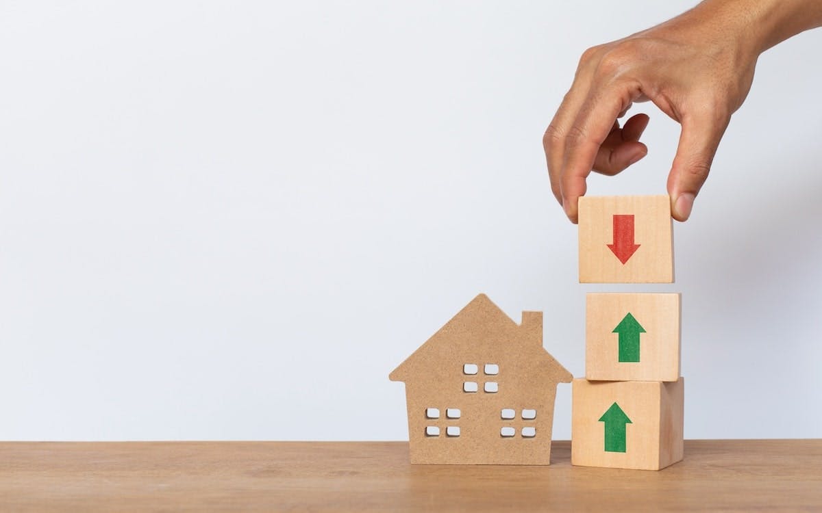 A hand places a wooden block with a down arrow on top of two blocks that have upward arrows. The stack of cubes stands next to a wooden model house.