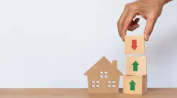 A hand places a wooden block with a down arrow on top of two blocks that have upward arrows. The stack of cubes stands next to a wooden model house.
