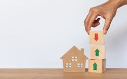A hand places a wooden block with a down arrow on top of two blocks that have upward arrows. The stack of cubes stands next to a wooden model house. A hand places a wooden block with a down arrow on top of two blocks that have upward arrows. The stack of cubes stands next to a wooden model house.