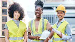 Three young women stand at a construction site wearing yellow vests