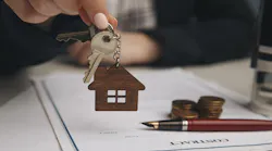 A real estate agent hands over a house key to a buyer. A contract with a pen on the desk indicates the contract was recently signed.