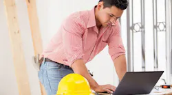 A young construction worker types on a laptop. A yellow hard hat sits off to the side.