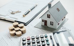 Shown is a table with a mortgage agreement, model home, calculator, and several stacks of coins. Shown is a table with a mortgage agreement, model home, calculator, and several stacks of coins.