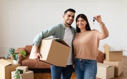 A couple stands in their new house surrounded by boxes from moving house. One person carries a cardboard box, while the other holds up the house key. A couple stands in their new house surrounded by boxes from moving house. One person carries a cardboard box, while the other holds up the house key.