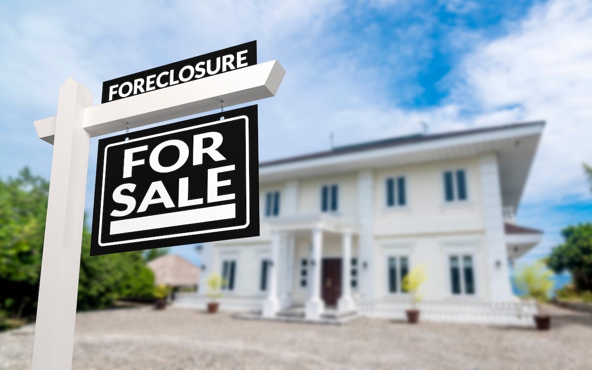 A black 'Foreclosure' sign is posted in the front yard of a large white house