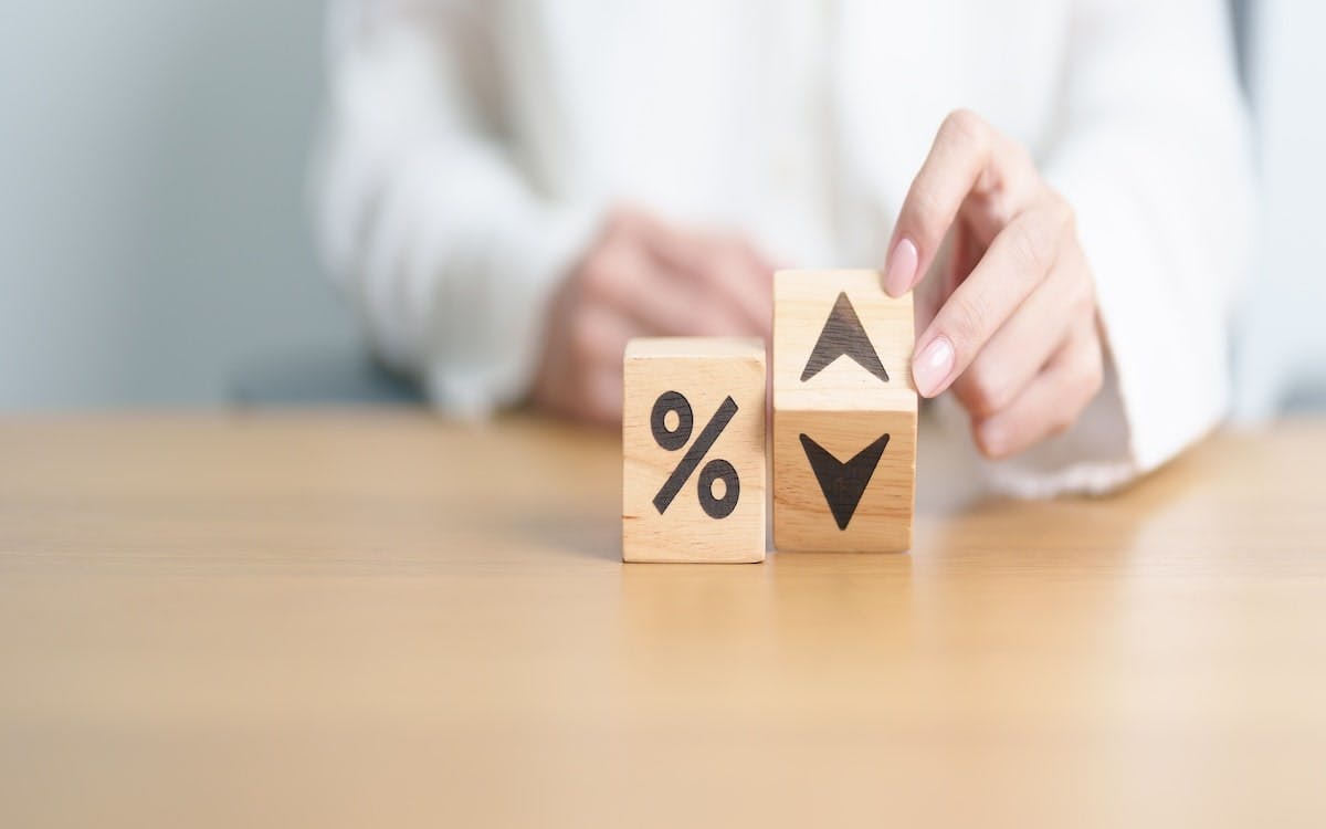 Two wooden blocks on a table. One block has a percentage symbol, the other has an up and down arrow. A hand moves the second block to show the 'up' arrow, symbolizing an increase in rates.