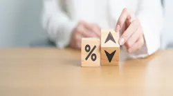 Two wooden blocks on a table. One block has a percentage symbol, the other has an up and down arrow. A hand moves the second block to show the 'up' arrow, symbolizing an increase in rates.