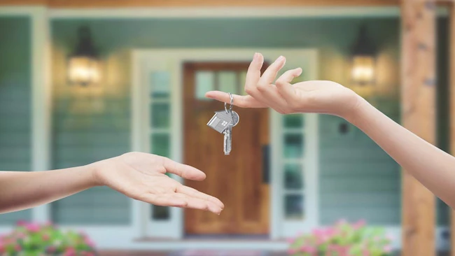 One individual hands a set of keys to another individual. A home sits in the background.