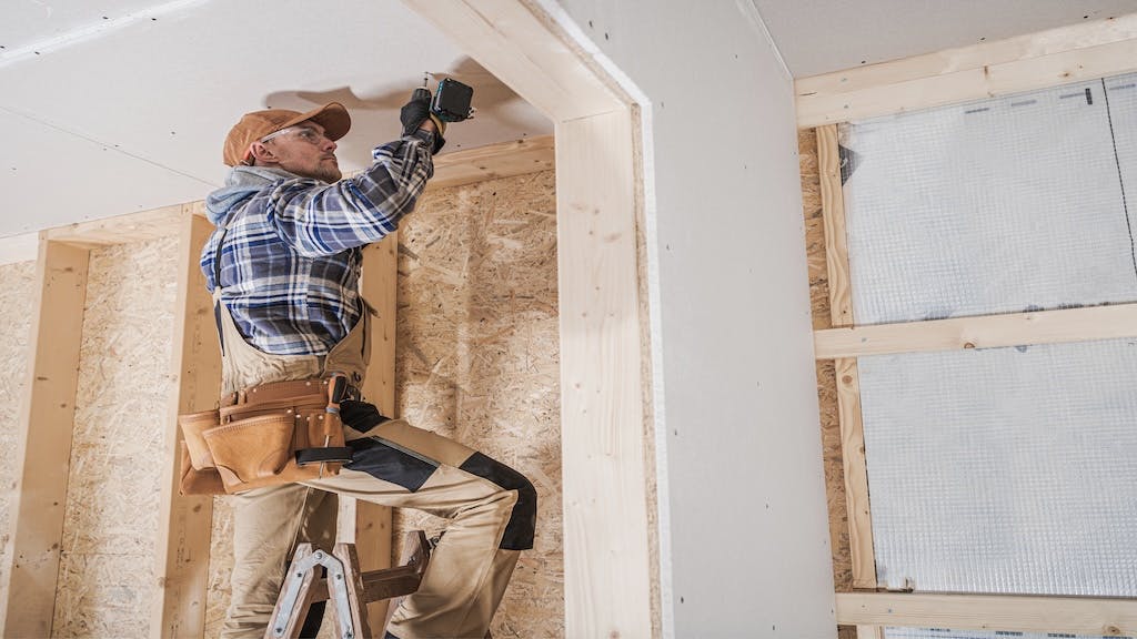 A construction worker stands on a ladder to get closer to ceiling dry wall.
