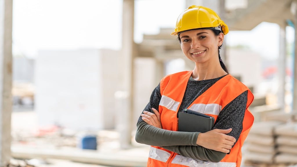 A young woman in a hard hat and orange vest poses in front of a construction site.