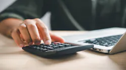 A homeowner sits at a desk typing on a laptop while going over home financing figures on a calculator.