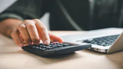 A homeowner sits at a desk typing on a laptop while going over home financing figures on a calculator.
