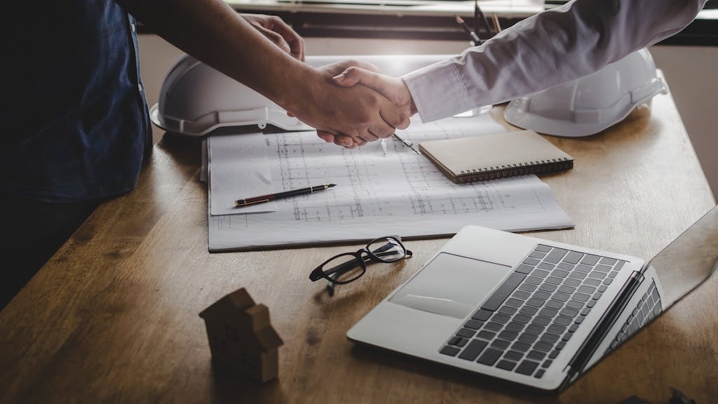 A handshake over a table that has a laptop, construction drawings, and two hard hats on it.
