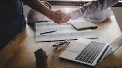 A handshake over a table that has a laptop, construction drawings, and two hard hats on it.
