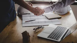 A handshake over a table that has a laptop, construction drawings, and two hard hats on it.