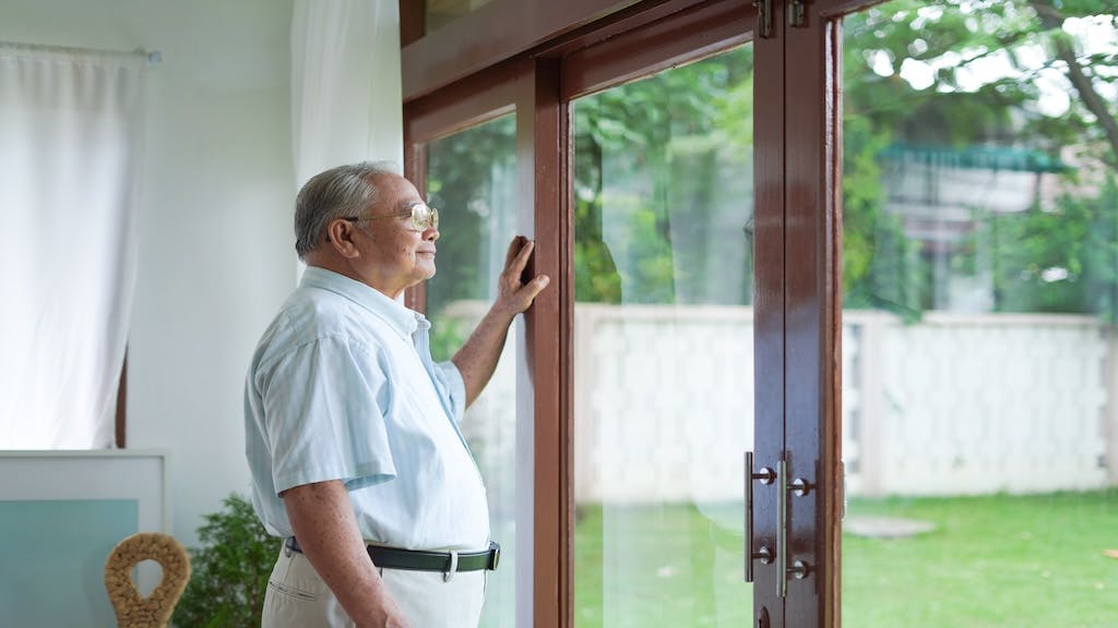 An elderly man looks out the window of his home to the backyard.