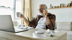 A Gen-X man talks on the phone while looking at a laptop and drinking coffee.