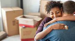 A woman hugs a man with a key to their new house in hand. Boxes behind them indicating that they are in the process of moving.