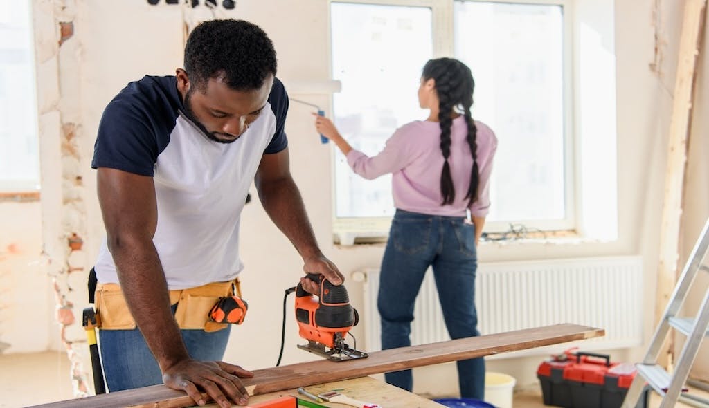 A man in the foreground uses a jig saw while a woman in the background paints the walls of a home they're remodeling to flip..