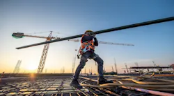 A construction worker walks over rebar carrying a beam on his shoulder.