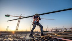 A construction worker walks over rebar carrying a beam on his shoulder. A construction worker walks over rebar carrying a beam on his shoulder.