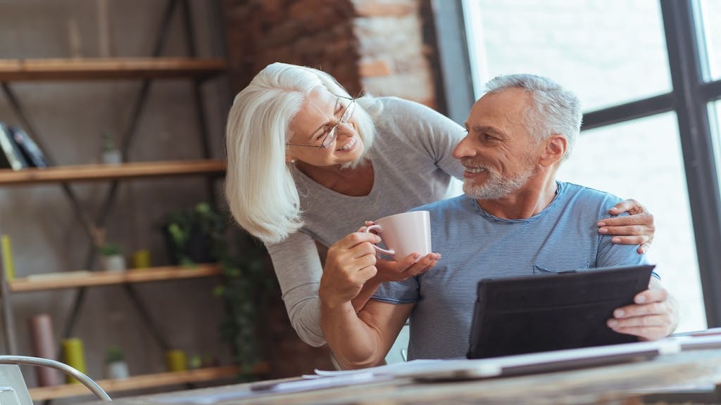 An elderly woman brings her husband a cup of coffee while he works at a laptop at a table in their home.