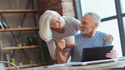 An elderly woman brings her husband a cup of coffee while he works at a laptop at a table in their home.