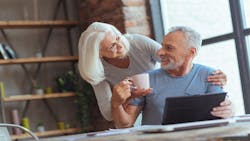 An elderly woman brings her husband a cup of coffee while he works at a laptop at a table in their home. An elderly woman brings her husband a cup of coffee while he works at a laptop at a table in their home.