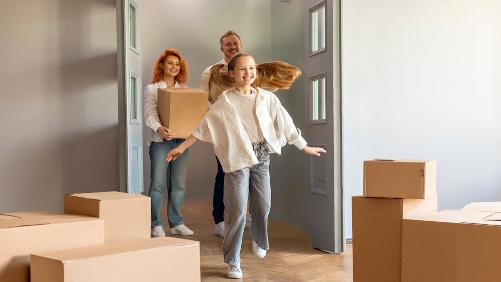 A family excitedly walks into their new home carrying moving boxes.