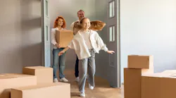 A family excitedly walks into their new home carrying moving boxes.