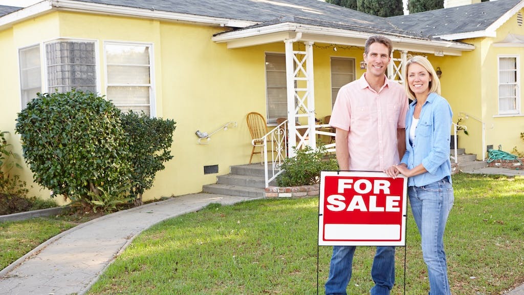Homeowners stand in front of their home with a 'For Sale' sign