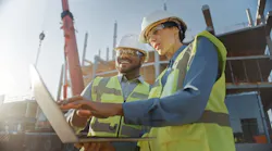 Two constructions work at a laptop with construction equipment behind them