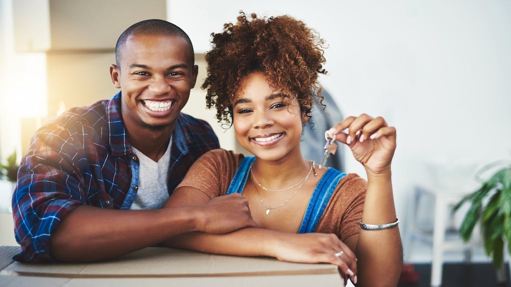 A young couple leans on moving boxes while holding up a set of house keys