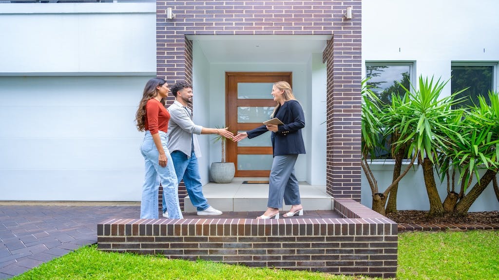 A young couple shakes hands with a real estate agent outside of their newly purchased home.