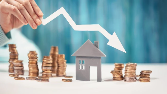 Coins stacked around a model home with an arrow pointing downward indicate decreased home prices.