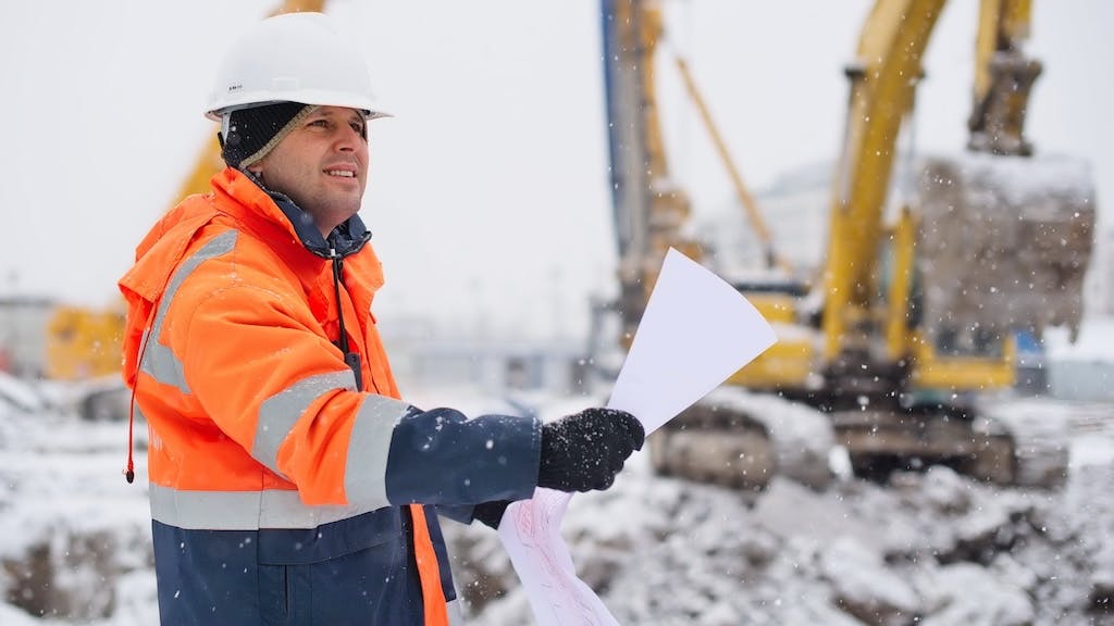 A construction worker stands outside in the snow looking over plans.