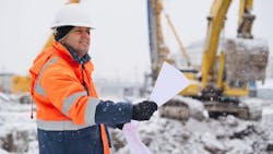 A construction worker stands outside in the snow looking over plans. A construction worker stands outside in the snow looking over plans.