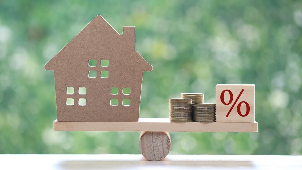 A model home sits on one side of a balancing beam while coins and a block with a percentage symbol sit on the other.