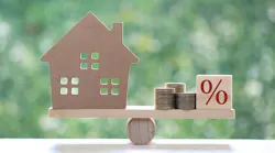 A model home sits on one side of a balancing beam while coins and a block with a percentage symbol sit on the other.