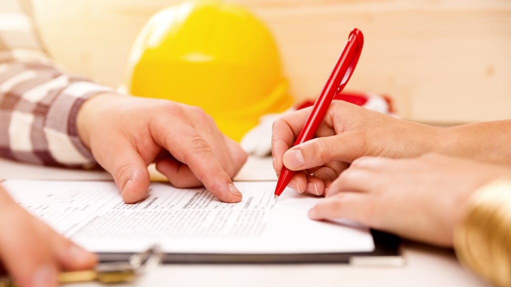 A closeup of a hand signing a document with a yellow hard hat in the background