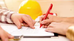 A closeup of a hand signing a document with a yellow hard hat in the background