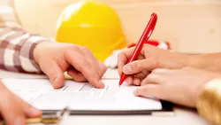A closeup of a hand signing a document with a yellow hard hat in the background A closeup of a hand signing a document with a yellow hard hat in the background