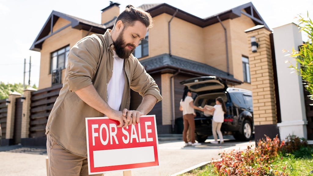A man sticks a 'For Sale' sign into his front lawn